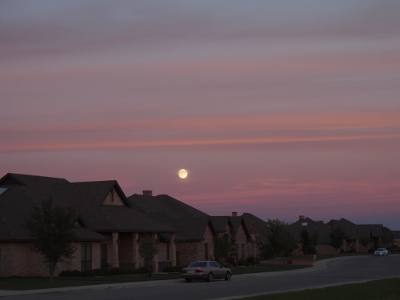 [What appears to be a full, bright-white moon sits just above the line of houses in a purplish sky with horizonal stripes of pink across it. There are some pinkish clouds across the upper part of the moon.]
